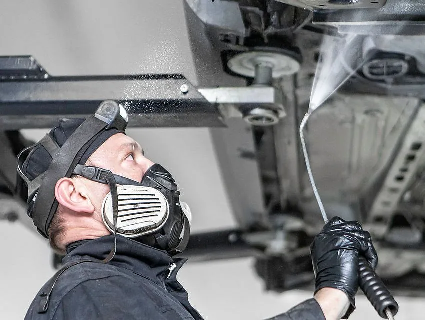 Mann med maske sprøyter behandling under bilen som er løftet i verksted.